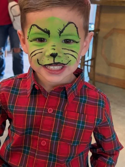 A young boy with face paint resembling a green cat, wearing a red plaid shirt, smiling in an indoor setting.