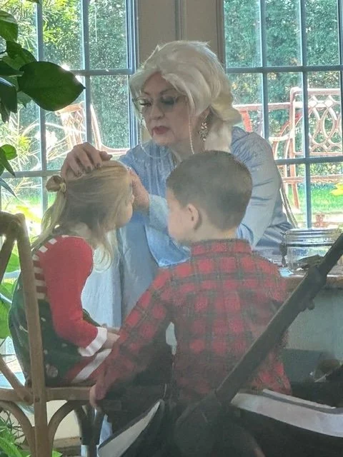 An older woman with white hair and glasses sitting at a table, gently brushing the hair of a young girl with blond hair, while a young boy in a red plaid shirt looks on. The scene takes place in a room with large windows and lush greenery outside.
