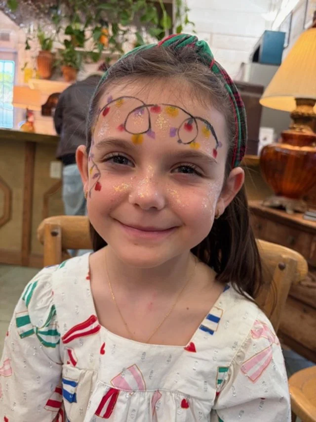 A young girl with face paint resembling Christmas lights on her forehead, cheeks, and nose, smiling indoors. She's wearing a white shirt with red and green patterns, a gold necklace, and a headband. The background includes plants, a lamp, and wooden furniture.