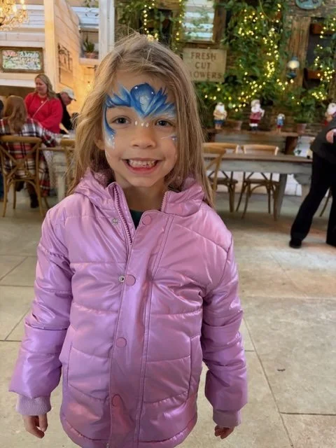 A young girl with face paint of a blue and white butterfly, smiling and wearing a pink puffer jacket in a decorated indoor setting.