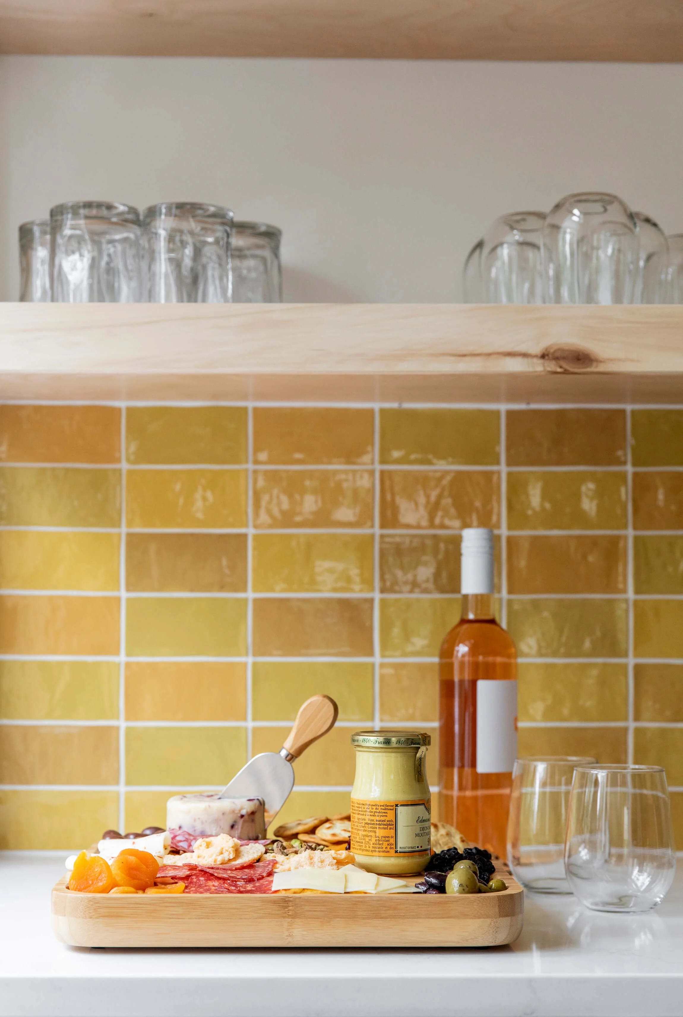Closing view of a wooden cheese and charcuterie platter with grapes, jam, slices of cheese, and dried apricots, on a white kitchen countertop with a yellow tiled backsplash.