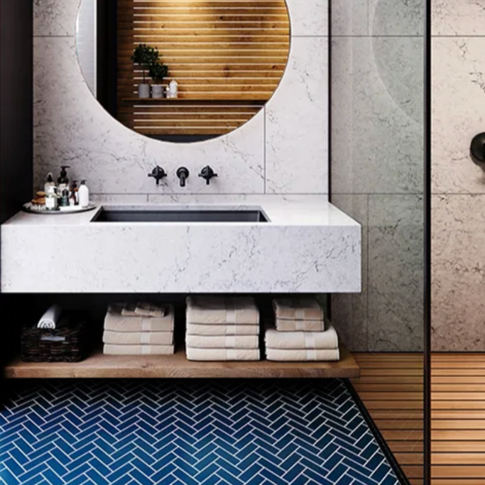 Modern bathroom with a white marble vanity, mounted black faucets, and a round mirror. Towels are stacked on a shelf below, with toiletries on the side. The wall has marble tiles, and there is a wooden accent wall with a small plant. The floor features a dark herringbone tile pattern.