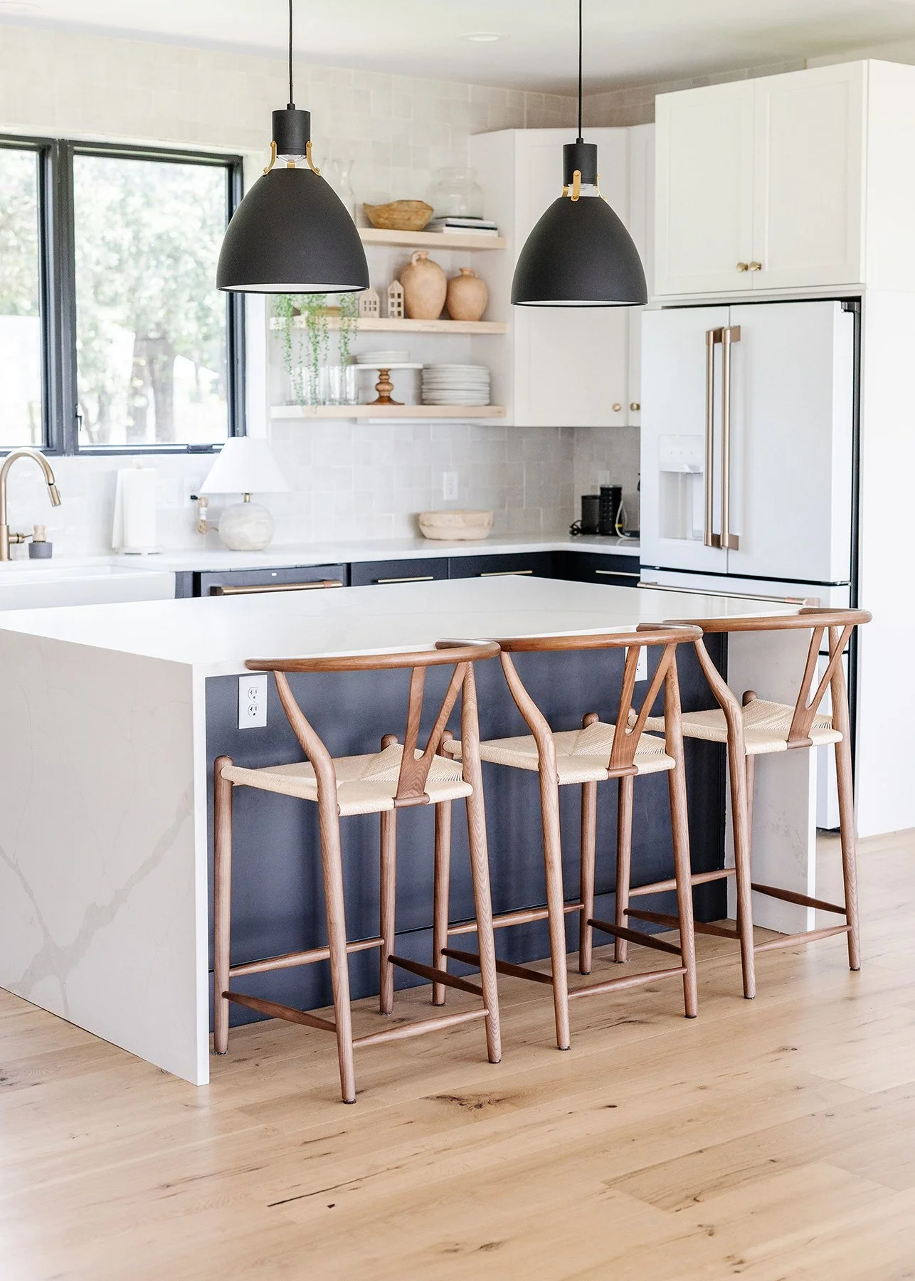 Modern kitchen with white countertops, navy cabinets, open shelving with pottery and dishes, large window, black pendant lights, three wooden barstools with woven seats at an island.