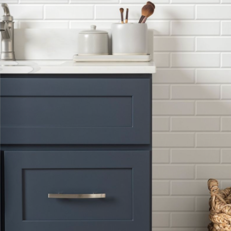 Close-up of a navy blue bathroom vanity with a white countertop, set against a white subway tile wall. Items on the counter include a gray container holding brushes, a small pitcher, and a light-colored round container.