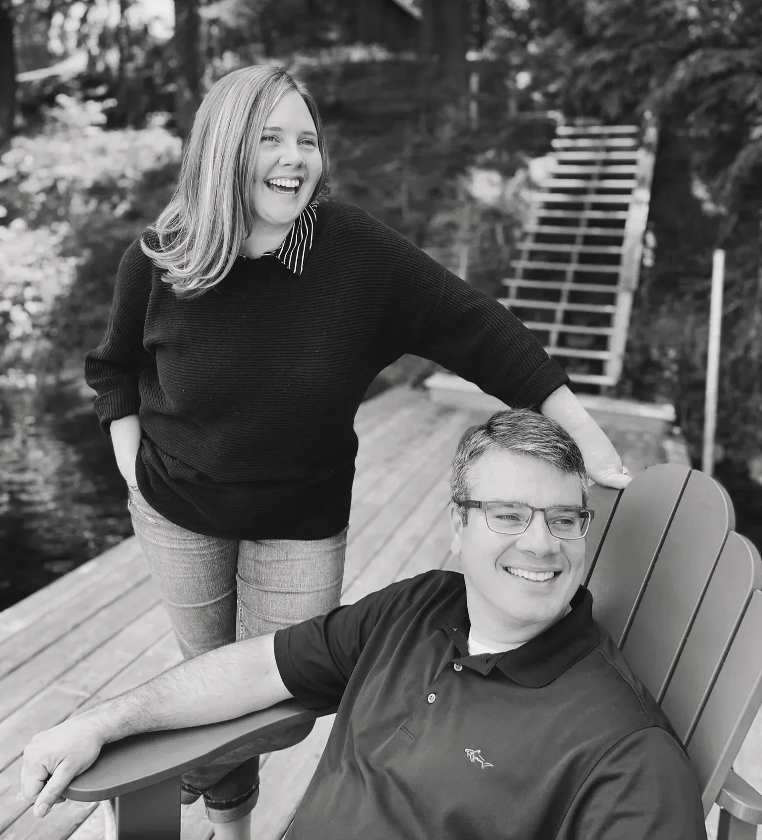A woman with shoulder-length hair and a striped shirt under a dark sweater is smiling and leaning on the shoulder of a man with glasses sitting in an outdoor chair. They are outdoors on a wooden deck with trees in the background.