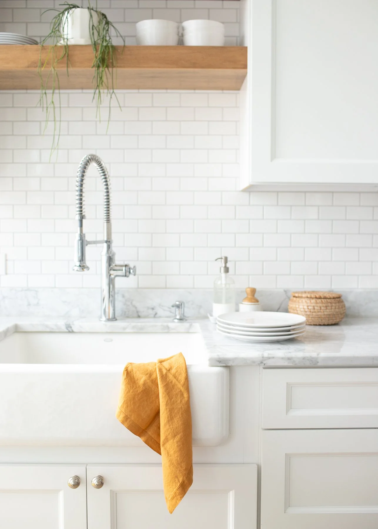 Kitchen sink with a yellow cloth hanging from it, white cabinets below, marble countertop, white tile backsplash, chrome faucet, soap dispenser, white plates, a woven basket, and a wooden shelf with bowls and a plant.
