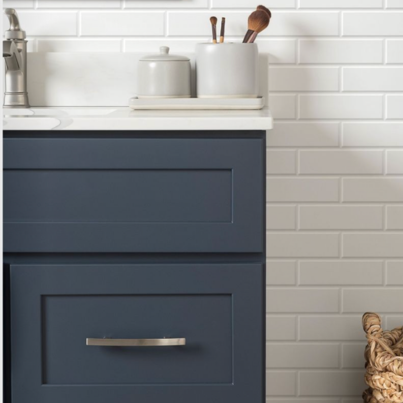 Close-up of a modern bathroom vanity with dark blue cabinetry, white countertop, and white ceramic container holding makeup brushes, against a white subway tile wall.