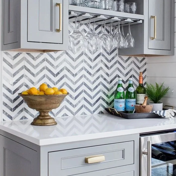 Kitchen with light gray cabinets, a white countertop, and a backsplash with a gray and white zigzag pattern. There are hanging wine glasses, a wooden bowl filled with lemons, bottles of sparkling water, a bottle of wine, a small potted plant, and a tray on the countertop.