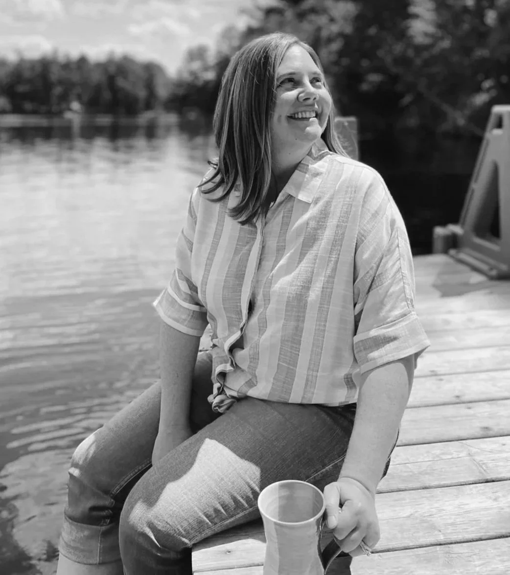 A woman smiling and sitting on a dock near a body of water, holding a mug, with trees in the background.