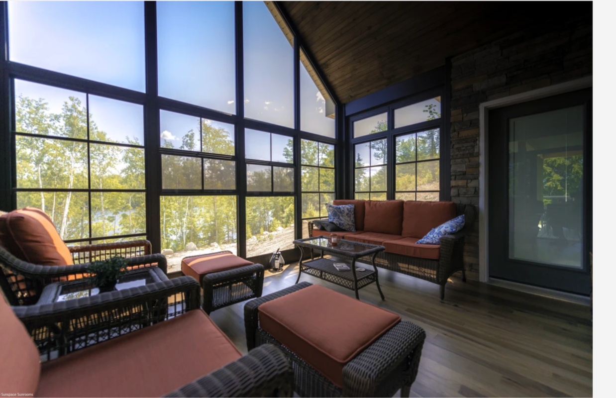 Sunroom with wicker furniture, orange cushions, and large windows showing a view of trees and a blue sky.