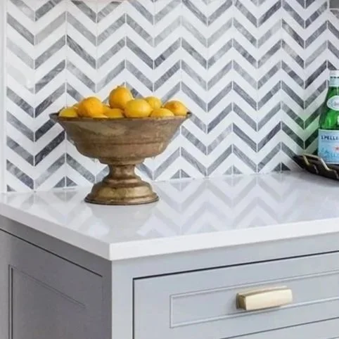 A wooden bowl filled with lemons sitting on a white countertop in front of a chevron-patterned gray and white tile wall. To the right, there's a tray with a green bottle and a glass.