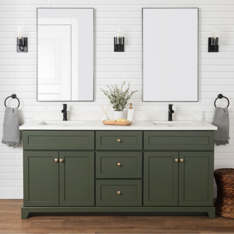 A bathroom double vanity with a green cabinet, two white sinks, black faucets, and two large mirrors on a white brick wall. There are black wall sconces with candles, gray towels on black rings, a basket, and a decorative tray with plants and toiletries.
