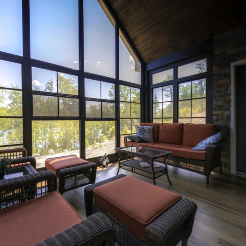 Sunroom with large floor-to-ceiling windows, wicker furniture with orange cushions, and a view of a wooded landscape outside.