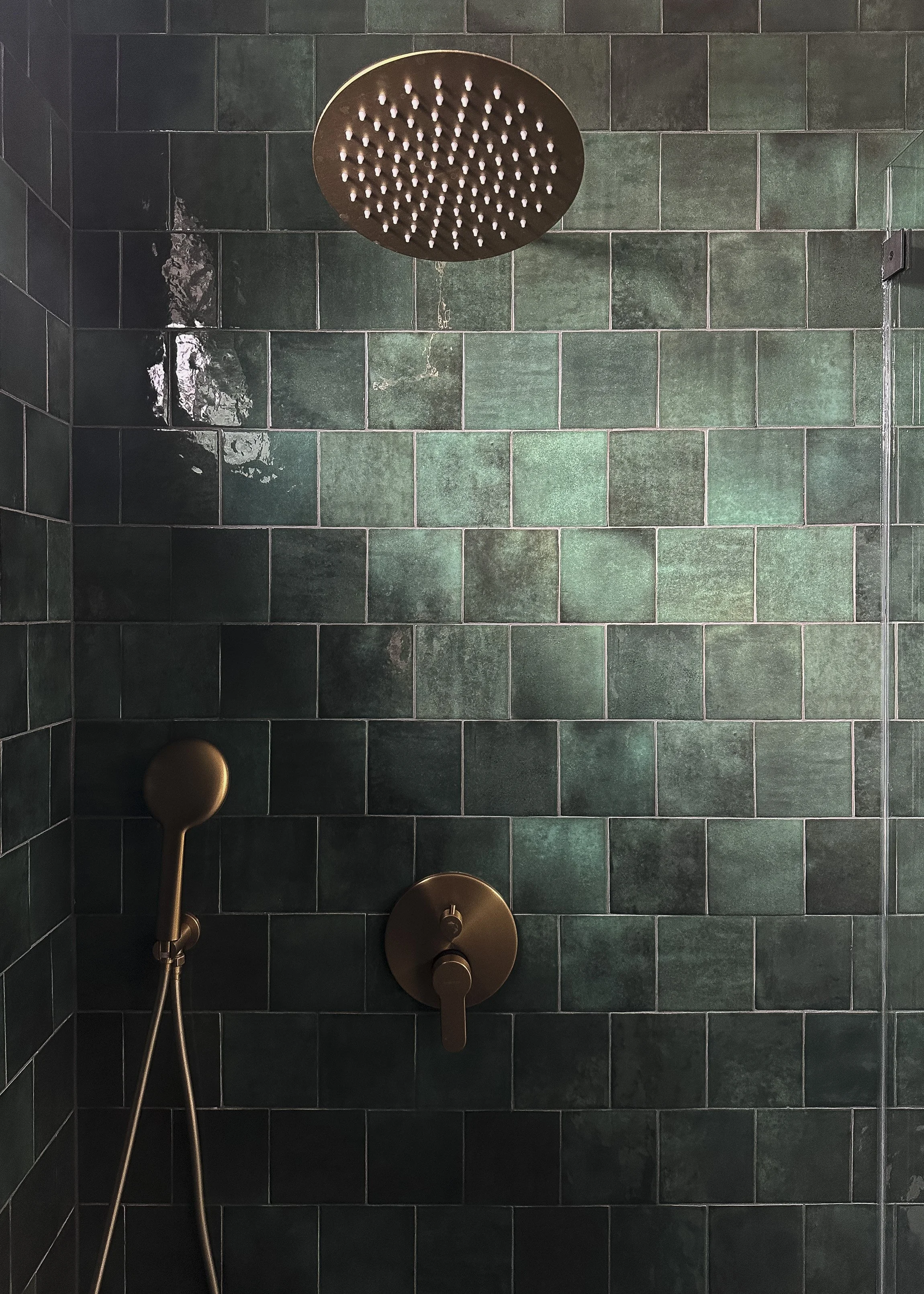 A modern shower with dark green tiles, a ceiling-mounted rain shower head, a handheld shower wand, and a single-handle control in a bronze finish.