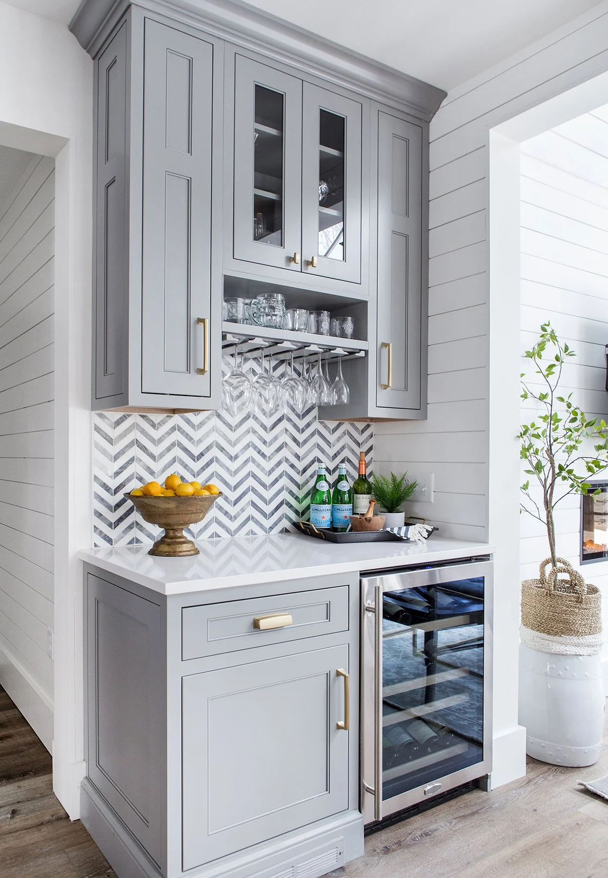 A modern kitchen with gray cabinets, a white countertop, a wine cooler, and a geometric tile backsplash, with glasses hanging above and plants nearby.