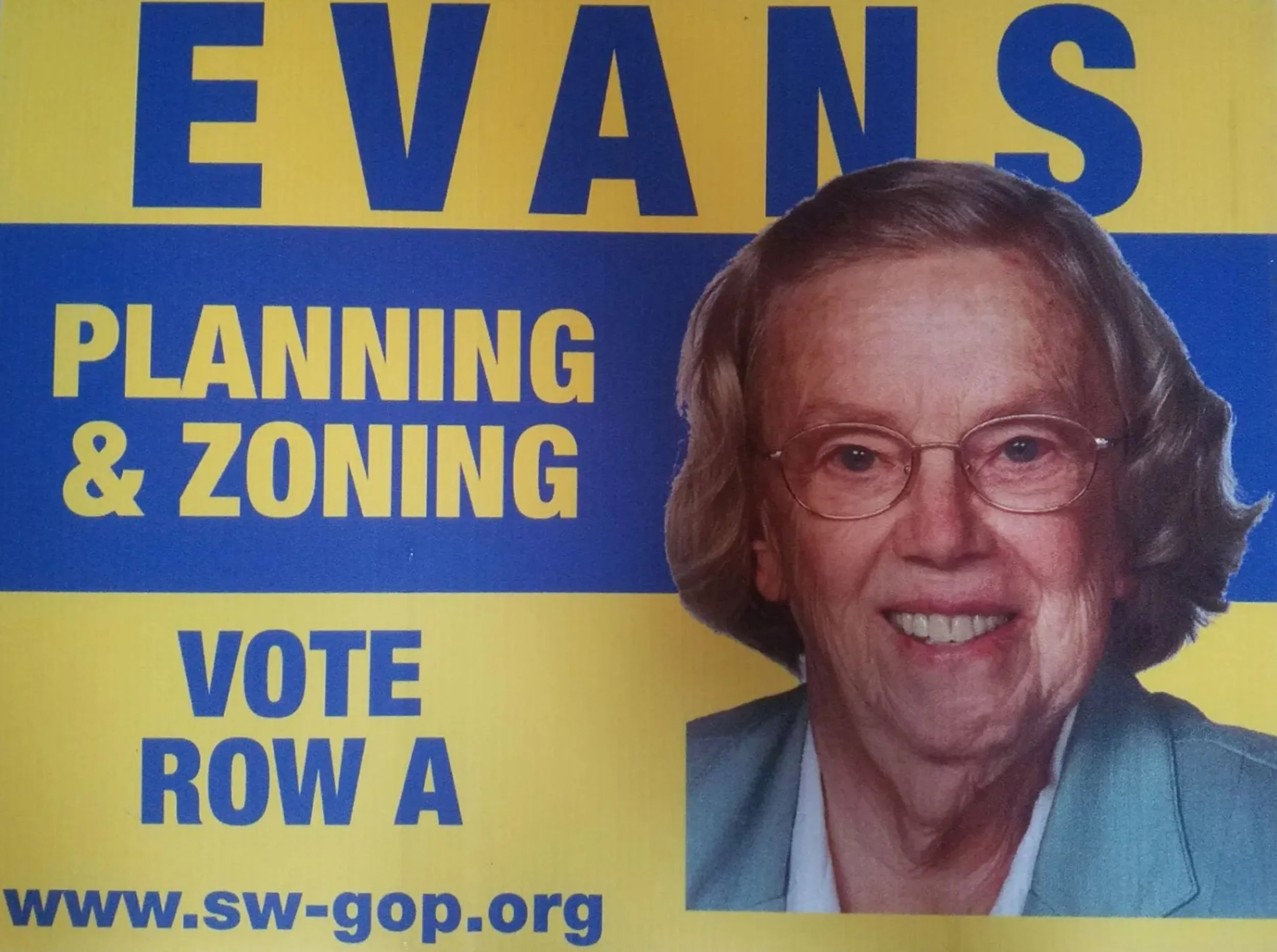 A woman with glasses and gray hair smiling, wearing a white shirt and blazer, in front of a yellow and blue sign that has the words "EVANS PLANNING & ZONING" and a web address.