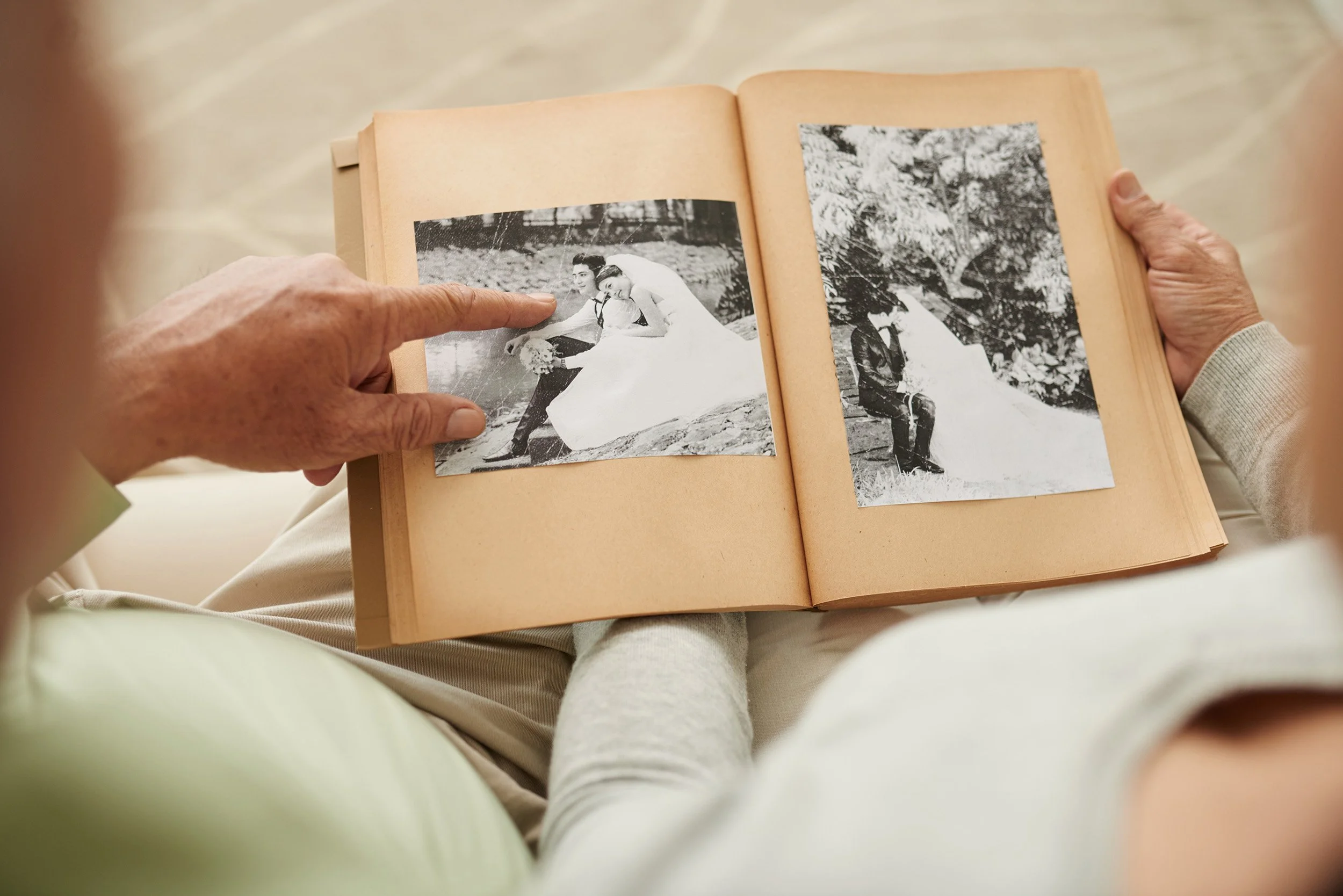 A person holding a photo album open while pointing at a black-and-white wedding photo of a bride and groom outdoors.