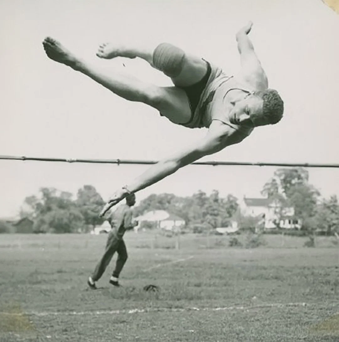 A black-and-white photo showing a man holding a pole with a large gymnast appearing to perform a flip in the air, creating an optical illusion of a giant in mid-air.