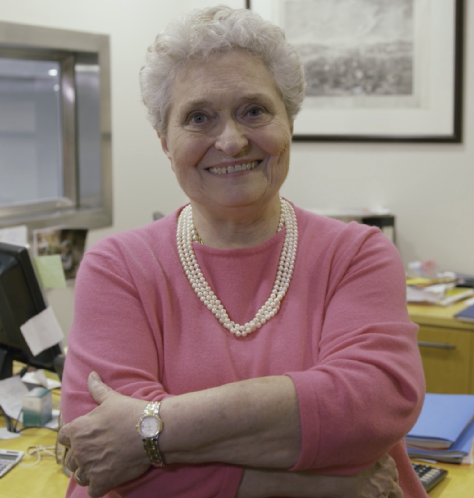 An elderly woman with short gray hair, smiling, wearing a pink sweater, a pearl necklace, and a watch, sitting at a desk in an office.