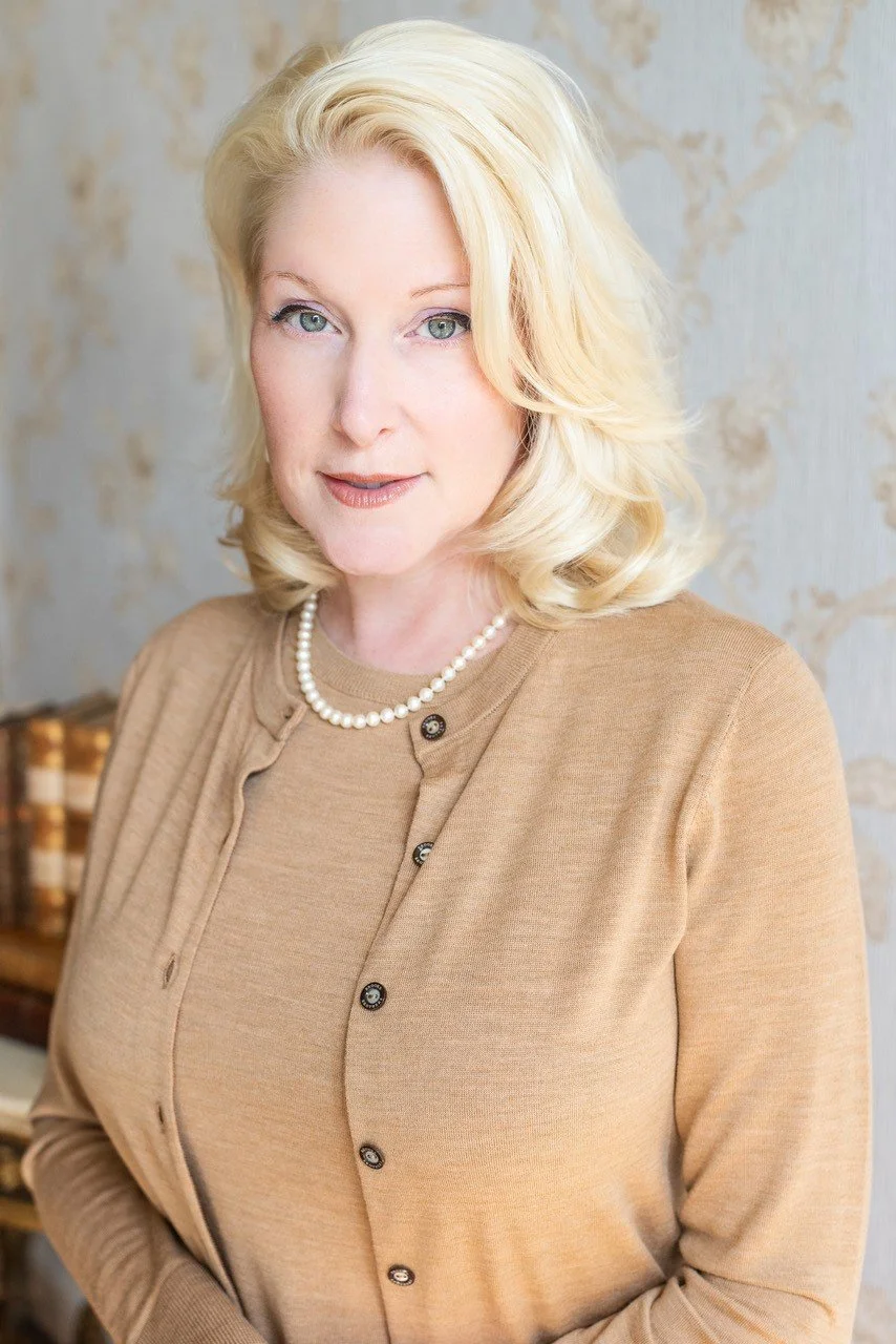 A woman with blonde hair wearing a beige cardigan and pearl necklace, standing indoors against a light textured wall.