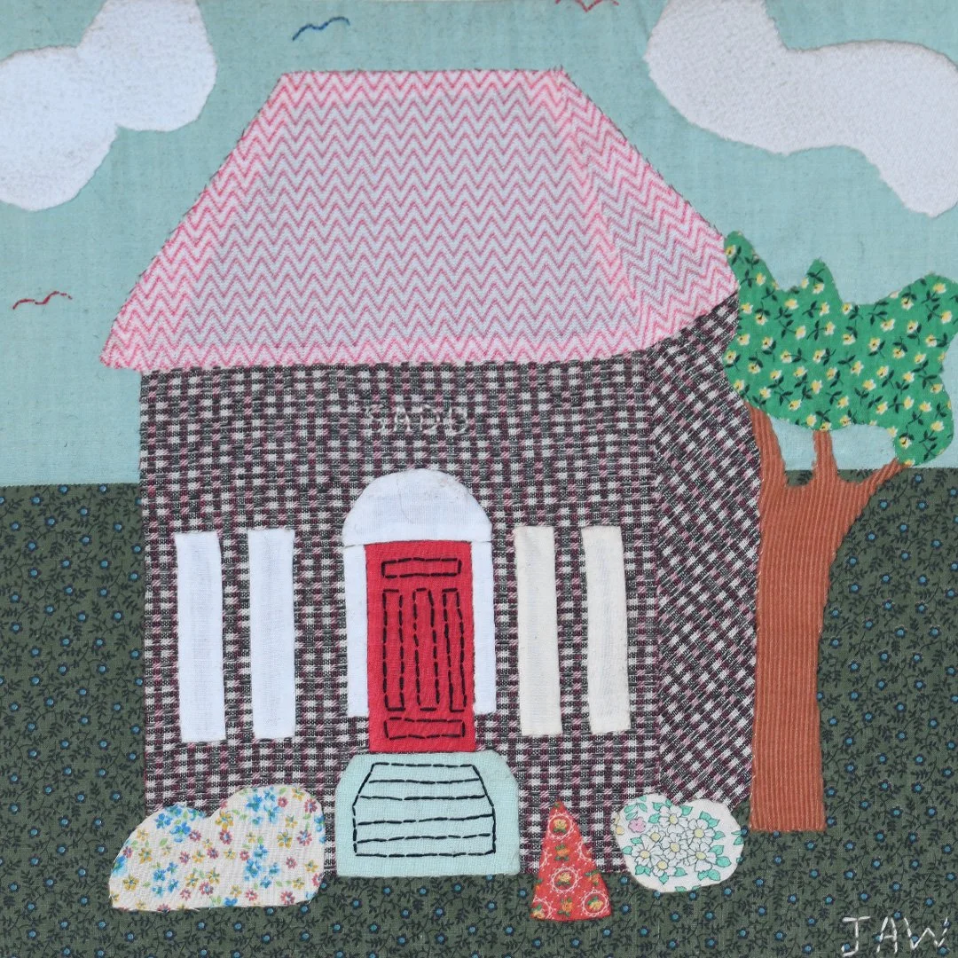 A fabric collage of a house with a pink and gray roof, a red door, white window frames, and a front staircase. Next to the house is a tree with green leaves and a brown trunk. There are bushes and decorative objects in front of the house, set against a sky background with clouds and birds.
