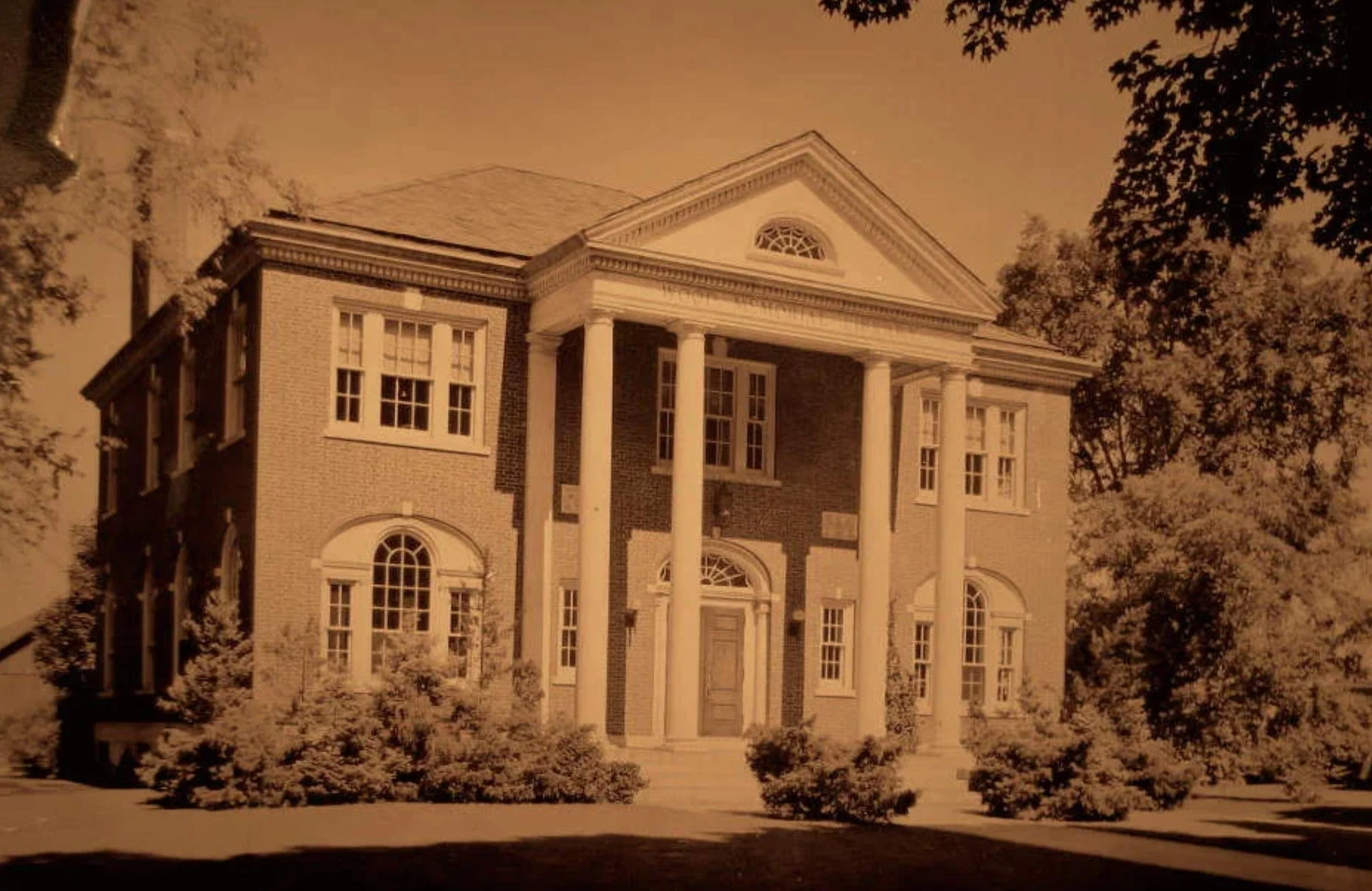 A large, elegant brick house with white columns at the entrance, surrounded by trees and bushes, with a cloudy sky overhead.