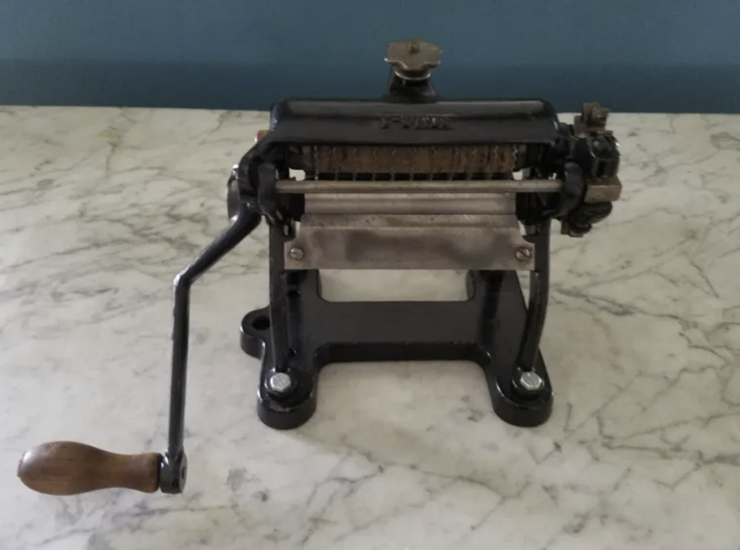 Vintage manual meat slicer with a wooden handle, metal frame, and cheese grater attachment, placed on a marble surface.