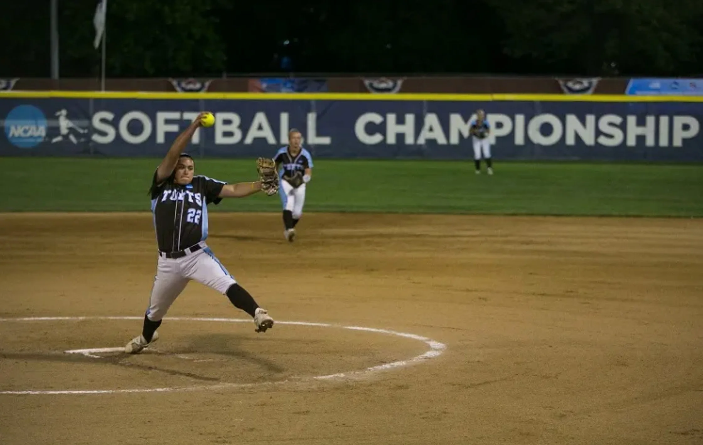 A female softball player in a black uniform with the number 22 is pitching a ball from the mound on a softball field during night time, with a large banner reading 'NCAA SOFTBALL CHAMPIONSHIP' in the background.
