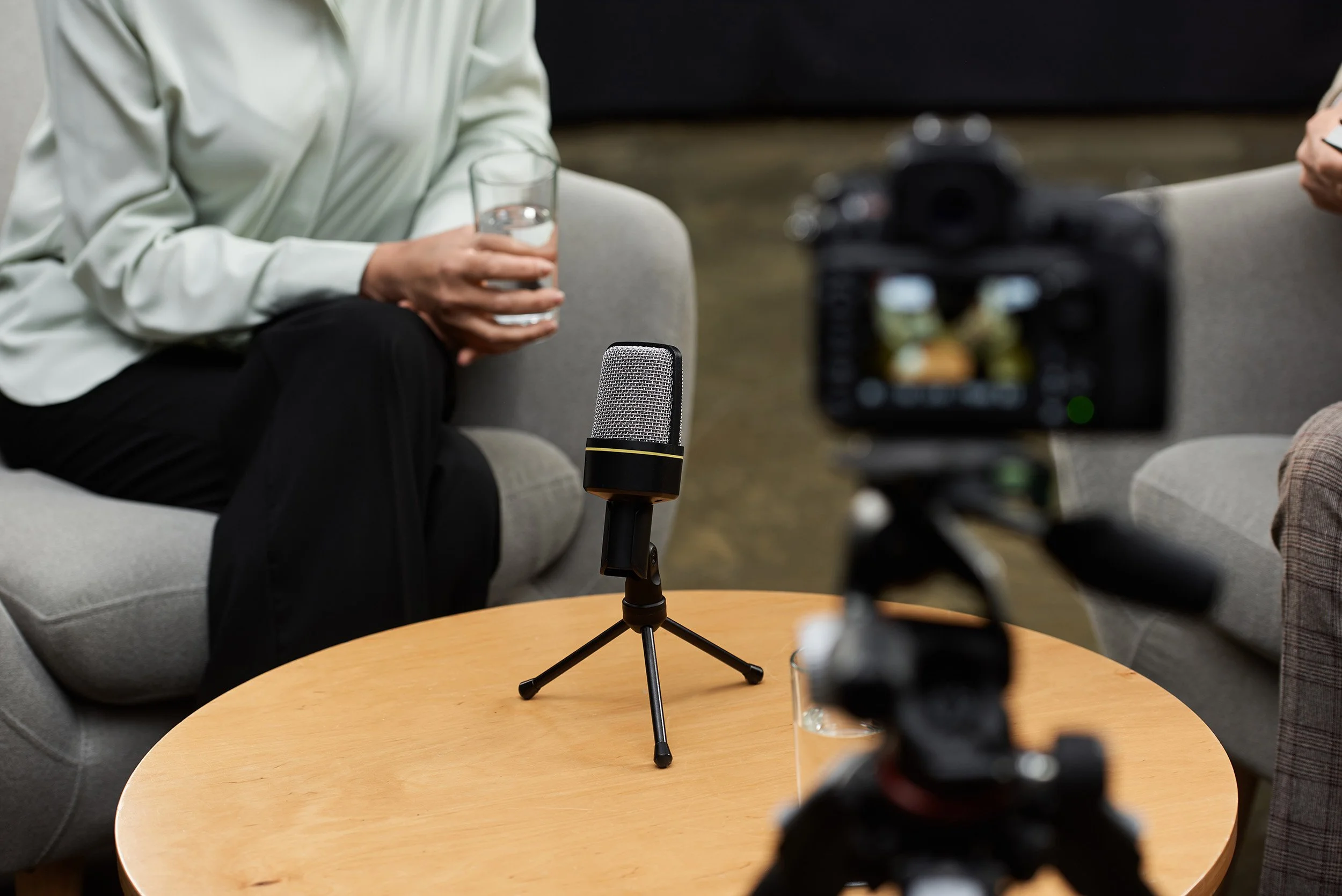Person sitting on a gray couch holding a glass of water, recording an interview with a microphone on a small wooden table between them, and a black camera positioned in front.