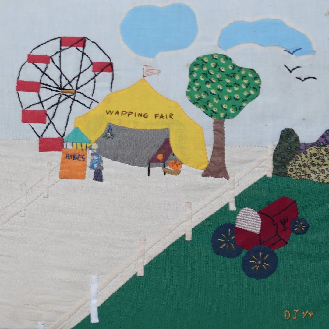 A colorful fabric illustration of a fairground with a ferris wheel, a yellow tent labeled 'WAPPING FAIR', a tree, a red ride, and a small wagon, with a cloudy sky and birds.