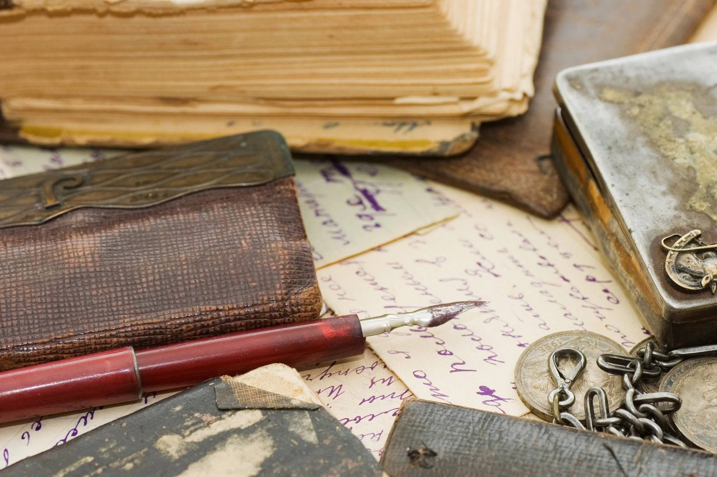 Close-up shot of vintage writing supplies including a fountain pen with a red barrel, worn leather-bound notebooks, handwritten letters with purple ink, small metal coins, and safety pins on a rustic surface.
