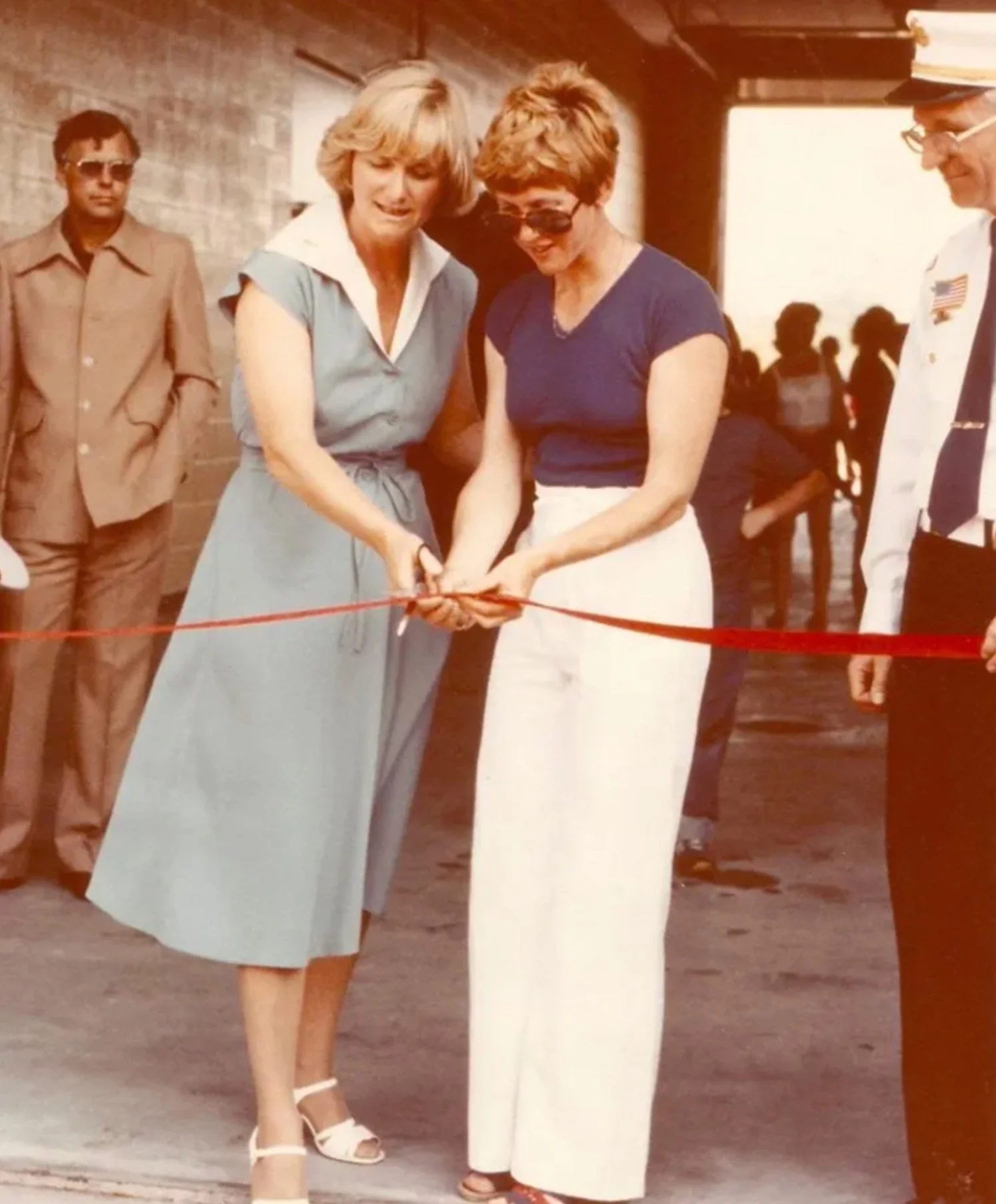 Two women are cutting a red ribbon at a door, with a man on the right and a man in sunglasses on the left observing them. They are dressed in casual clothing.