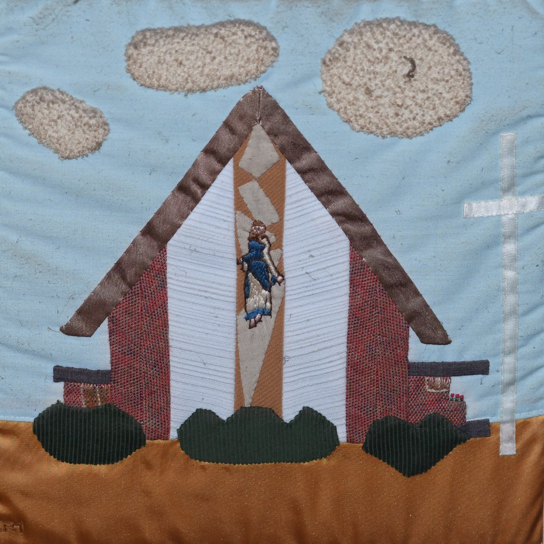 Embroidery of a church with a cross, clouds, bushes, and a religious figure at the center.