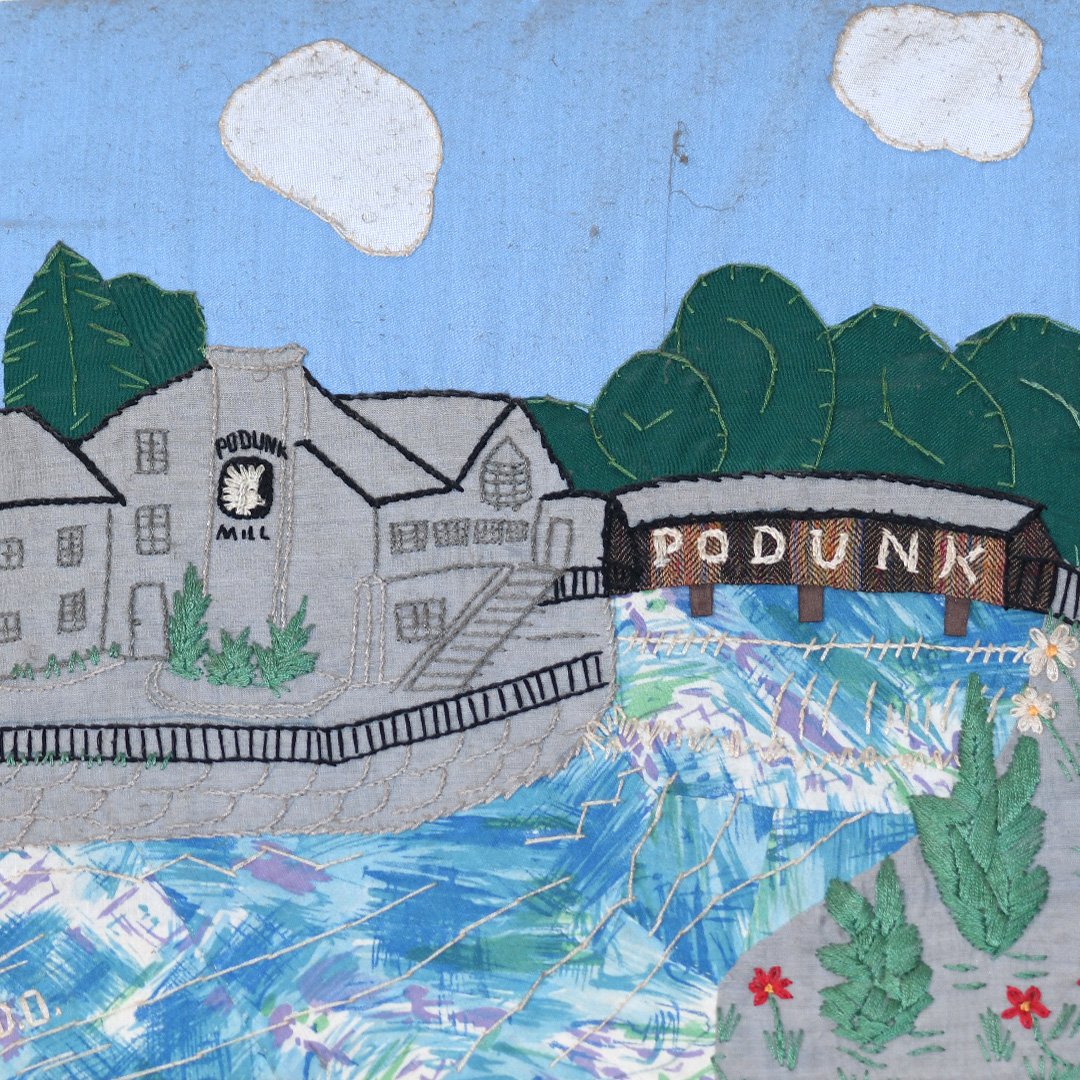 Embroidery of a landscape featuring a factory labeled 'Podunk Mill', a waterway, trees, clouds, and plants.