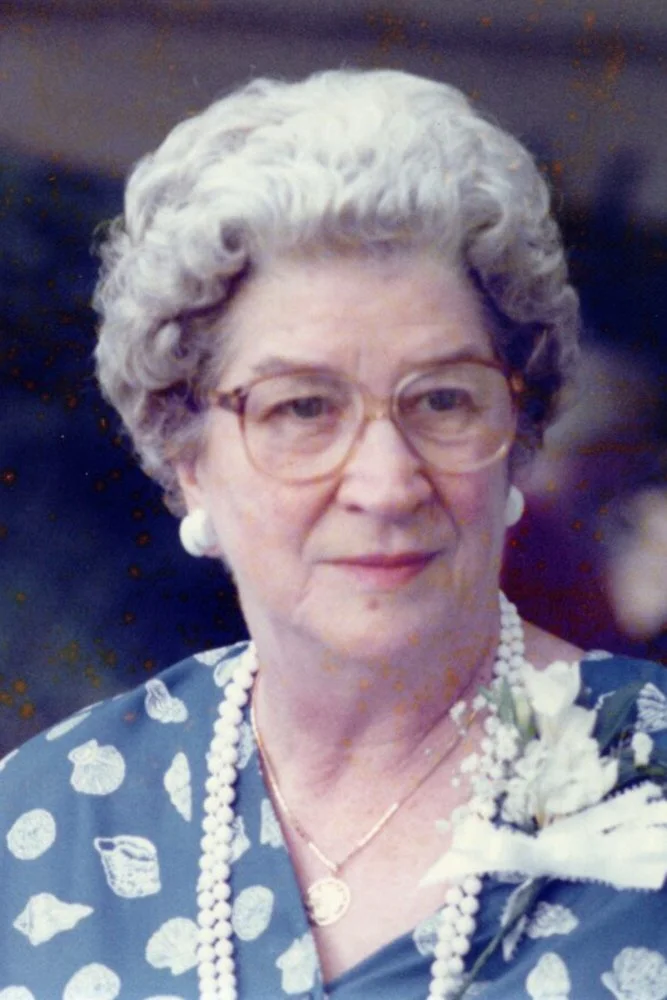 An elderly woman with short, curly gray hair, wearing glasses, pearl earrings, and a pearl necklace. She is dressed in a blue dress with white polka dots and is holding a white floral corsage.