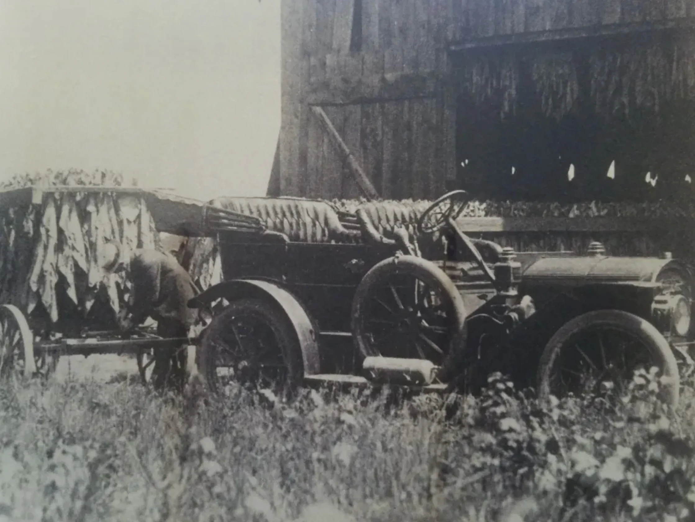 Black and white photograph of old farm equipment including a horse-drawn threshing machine, with a barn in the background.