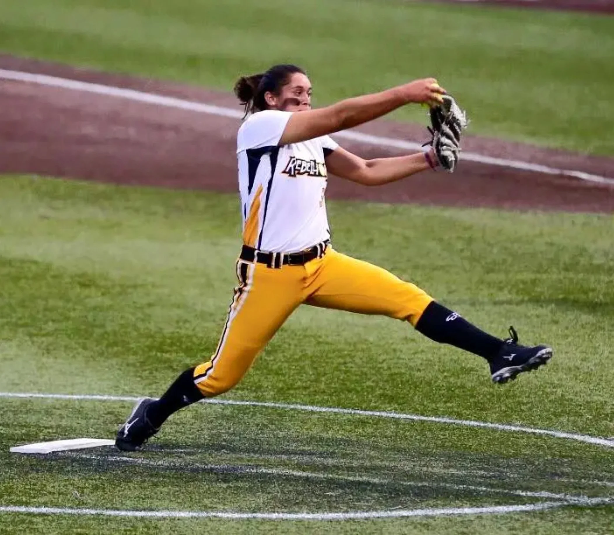 Female athlete in yellow athletic pants and a white jersey stretching on a grass field before a race.
