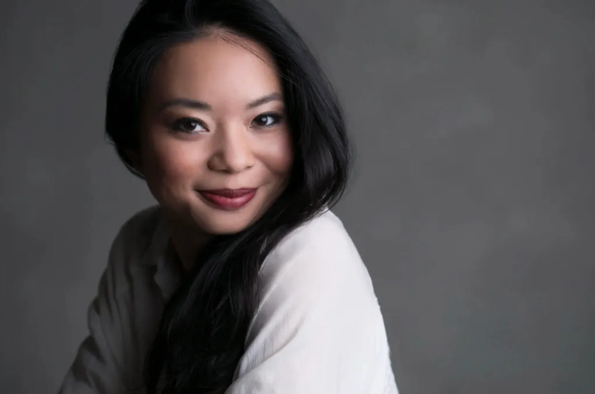 A woman with black hair in loose waves, wearing a white top, smiling, against a gray background.