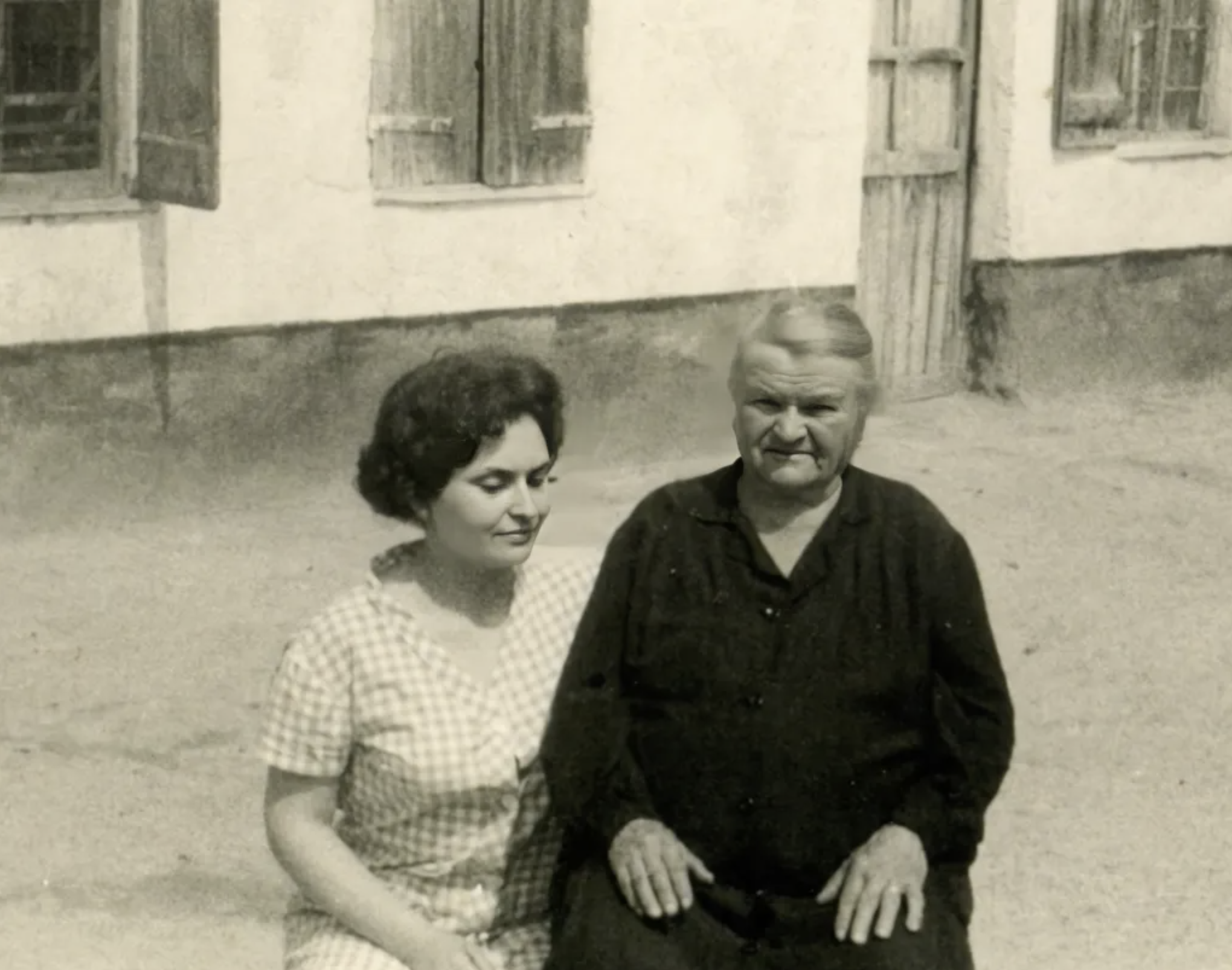 Black and white photograph of two people, a woman with dark hair and a man with light hair, sitting outside in front of a building with closed shuttered windows.