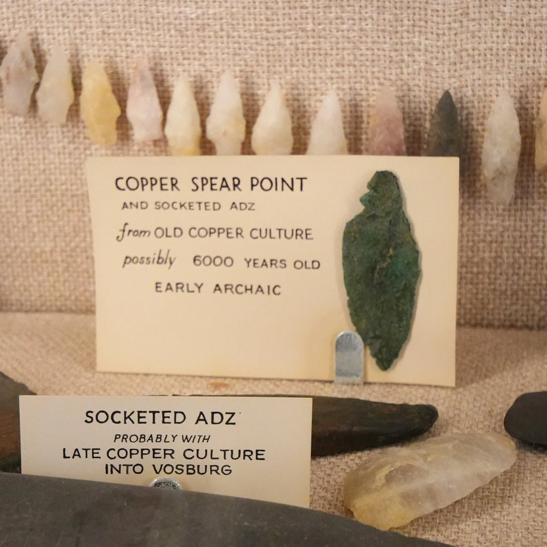 Close-up of a display featuring several small arrowhead-shaped stone points, labeled as copper spear points from ancient copper culture, with informational labels beneath.