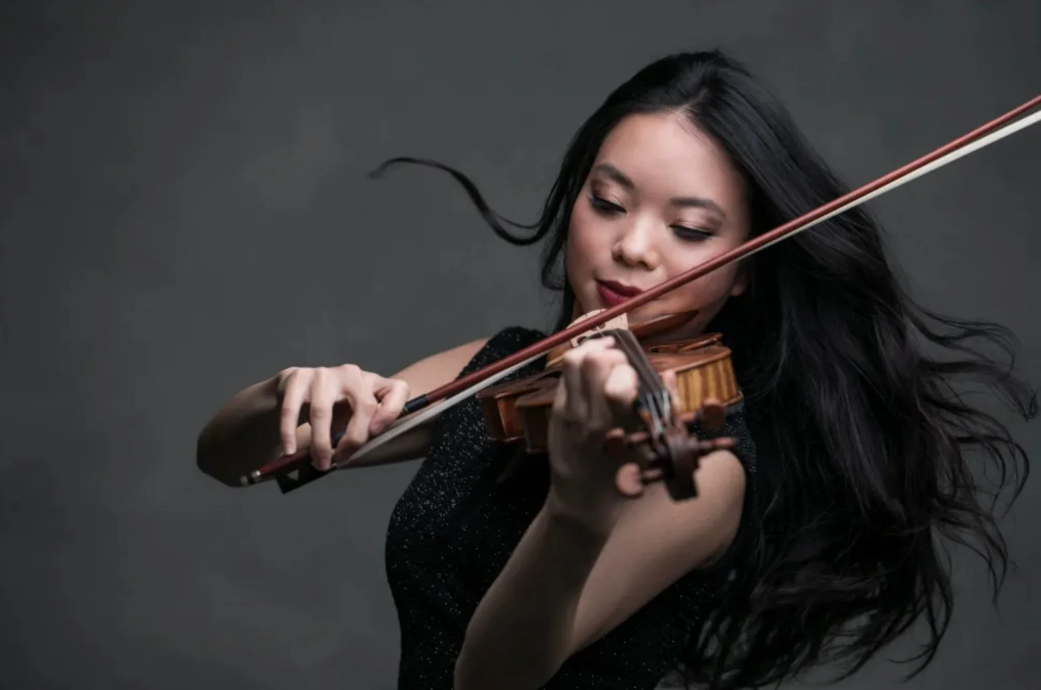 Woman with long black hair playing the violin against a gray background.