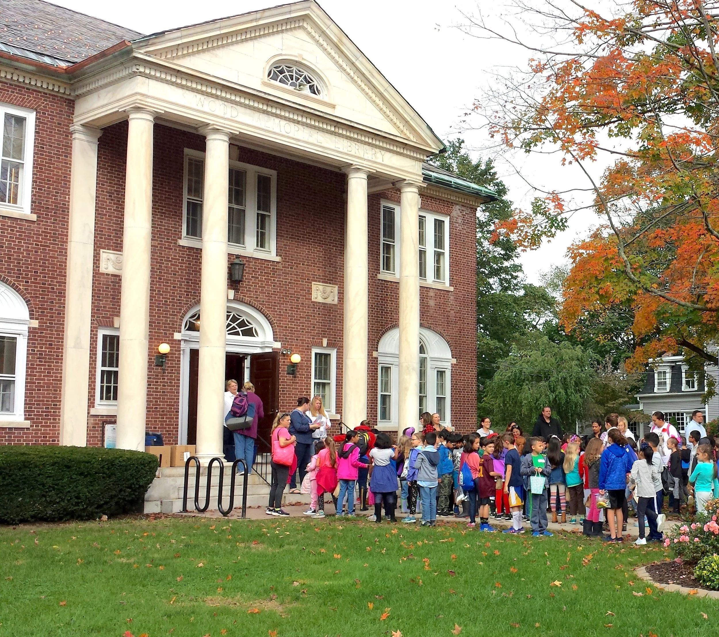 A school building with red brick walls and white columns, with a group of students gathered outside, some with backpacks, in front of a lawn with fallen autumn leaves, and a tree with orange leaves on the right side of the image.