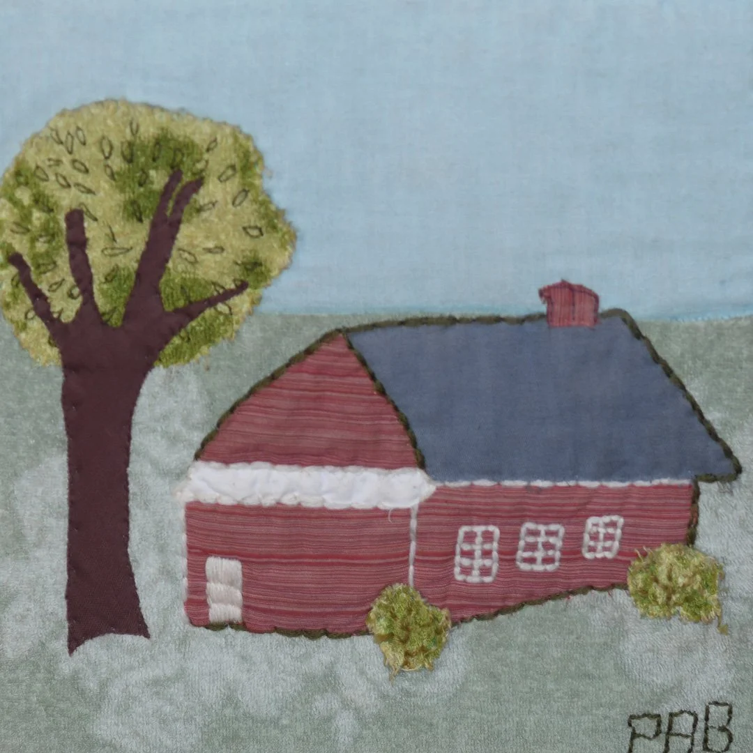 Embroidery of a house with a red roof, blue chimney, white window panes, green trees, and a blue sky.