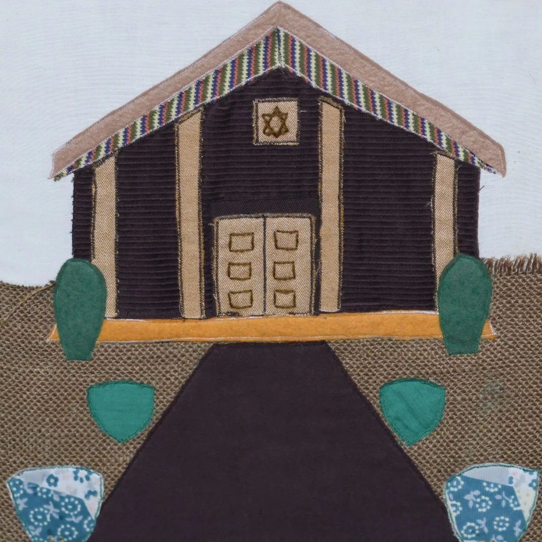 A fabric art depiction of a church with a black pathway leading to the door, green bushes on each side, colorful striped roof, and a small window above the door with a Star of David.