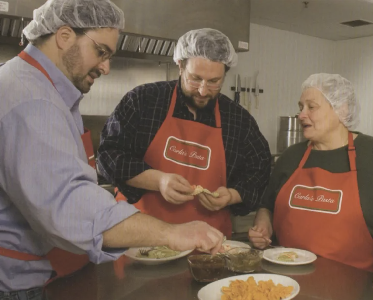 Three people wearing hairnets and aprons preparing food in a kitchen. They are focused on assembling dishes, with various bowls and plates of ingredients on the counter.