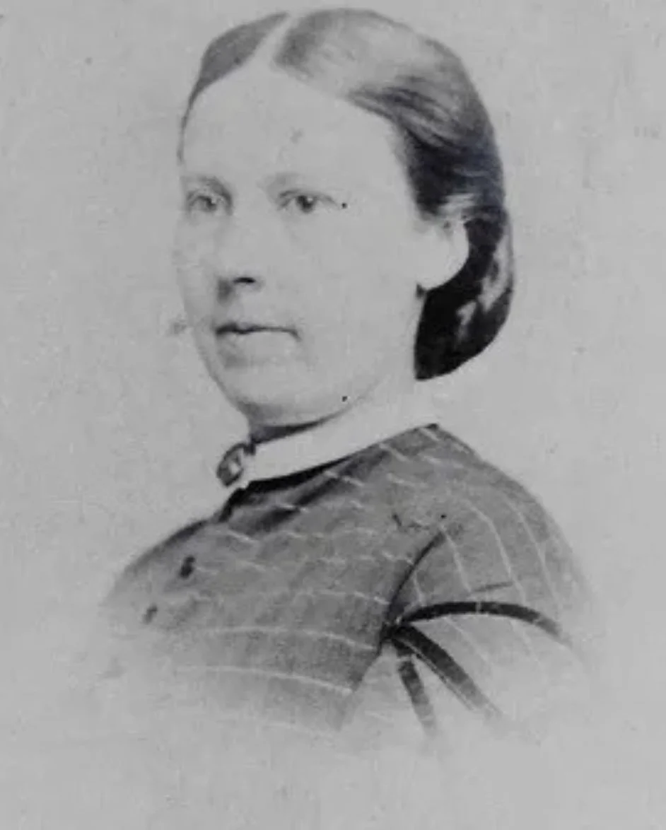 A black-and-white portrait of a woman with styled hair, wearing a checked dress and a collar with a bow, looking to her left.