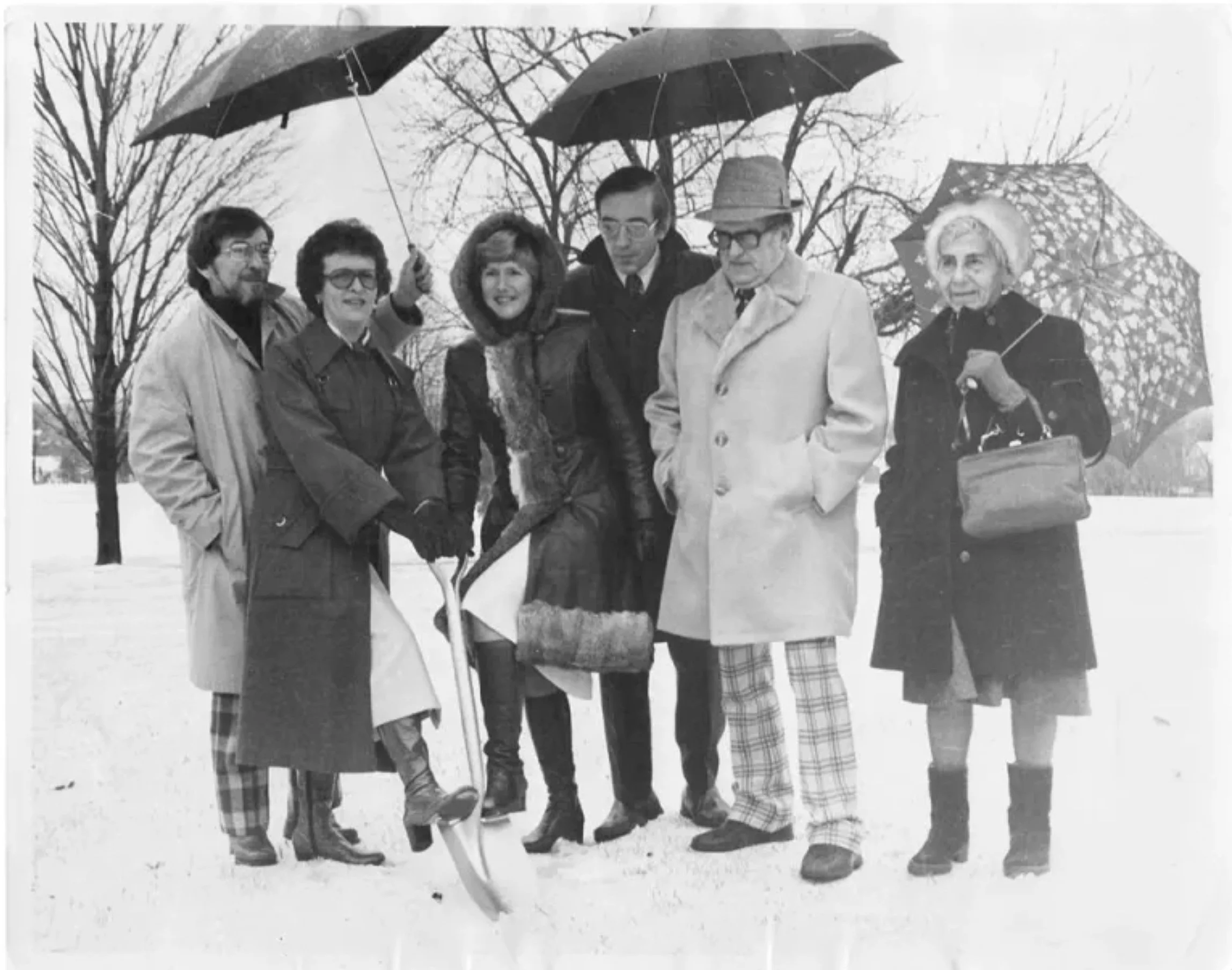 Black and white photo of six people standing outdoors in winter, dressed in warm clothing and holding umbrellas; three men and three women, with leafless trees and snow on the ground in the background.