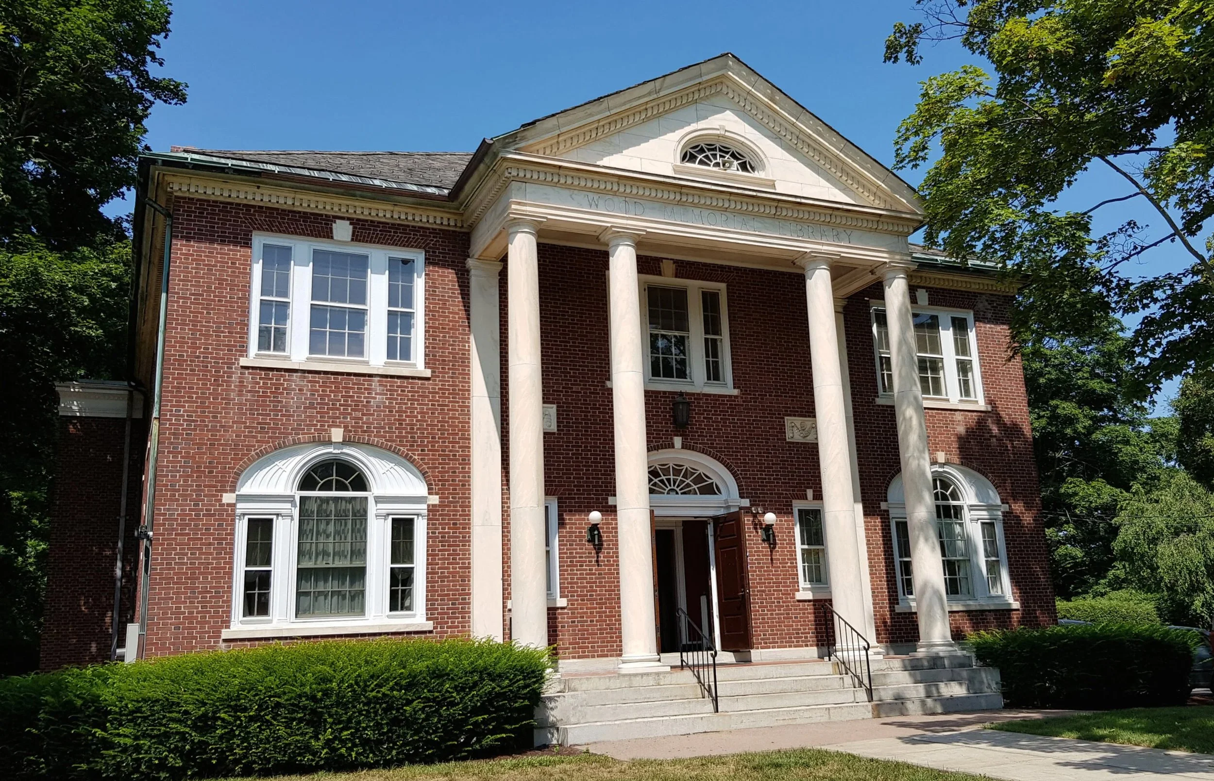 Red brick historic building with white columns and a triangular pediment, labeled "Wood Memorial Library," surrounded by greenery and trees, under a clear blue sky.