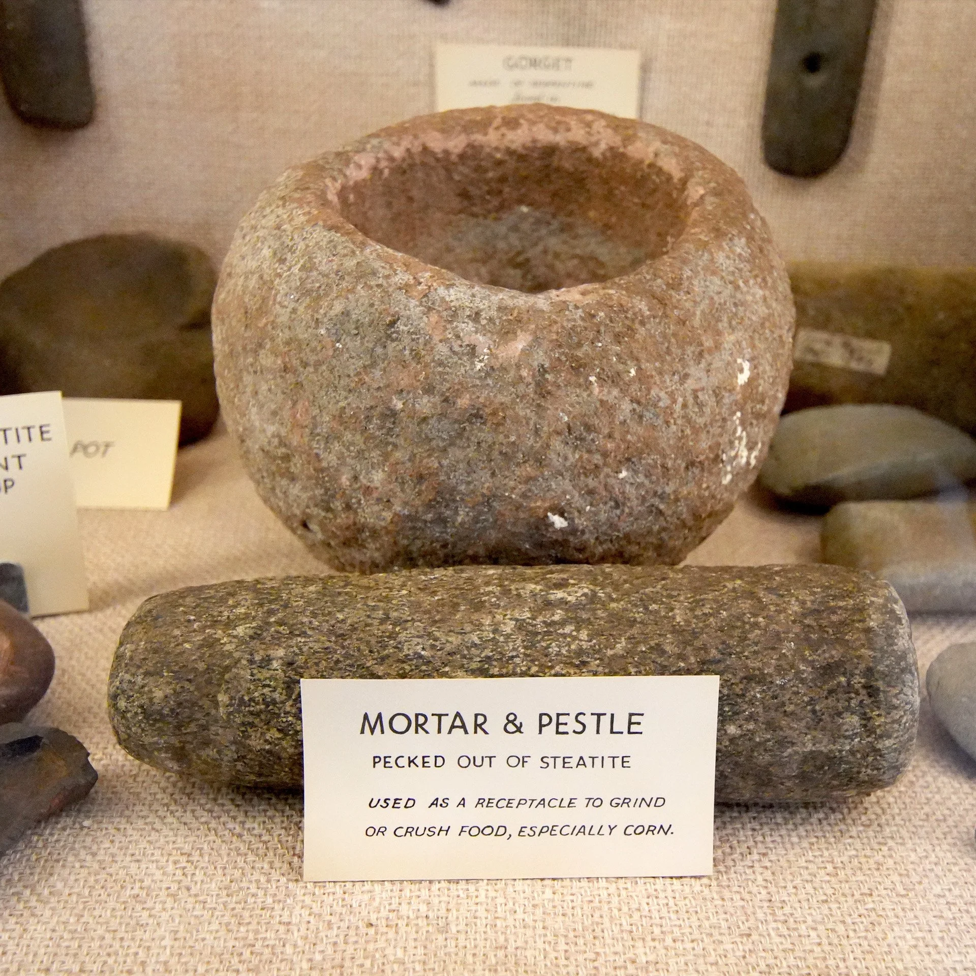 A rounded, rough-textured stone Mortar & Pestle with a small sign in front that reads: "MORTAR & PESTLE. Pecked out of Steatite. Used as a Receptacle to Grind or Crush Food, Especially Corn." In the background, there are other stones and a small part of a label visible.
