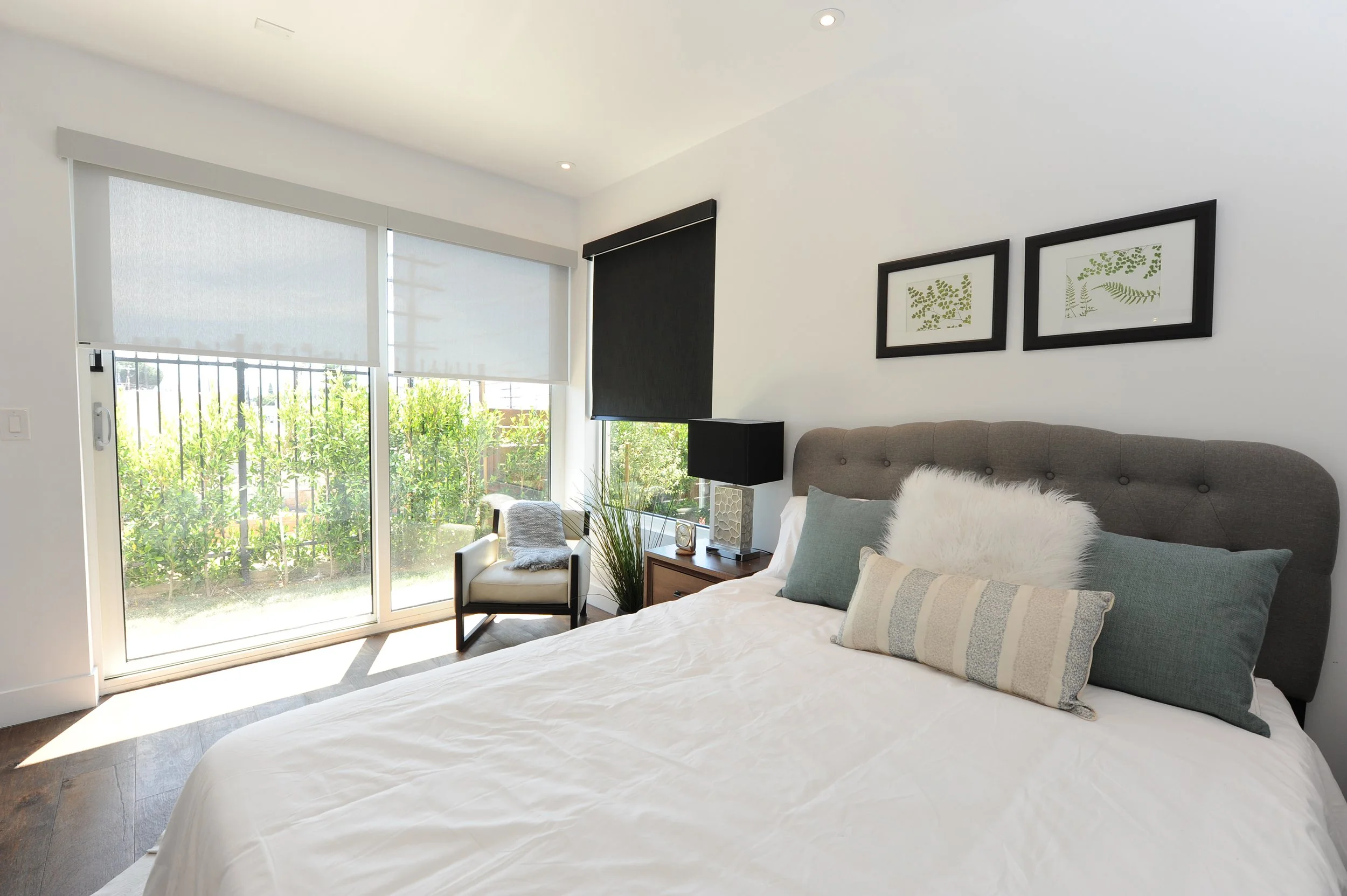 A bedroom with a large bed, gray headboard, several pillows, a nightstand with a lamp, and a window with white blinds and black blackout curtain. Outside, there is greenery and a fence.